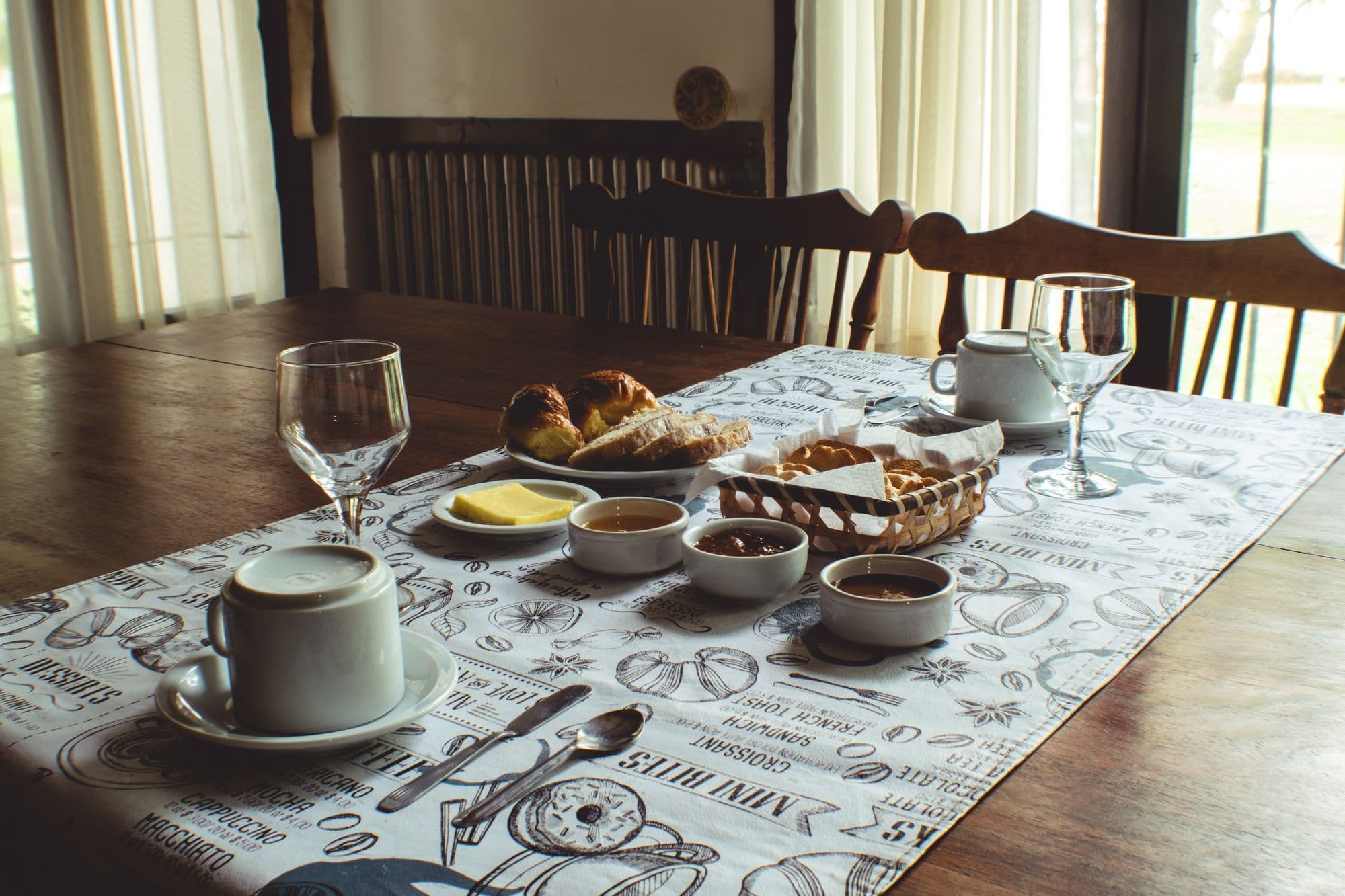 Een houten tafel bij het raam, gedekt voor het ontbijt: koffie, broodjes, boter en confituur