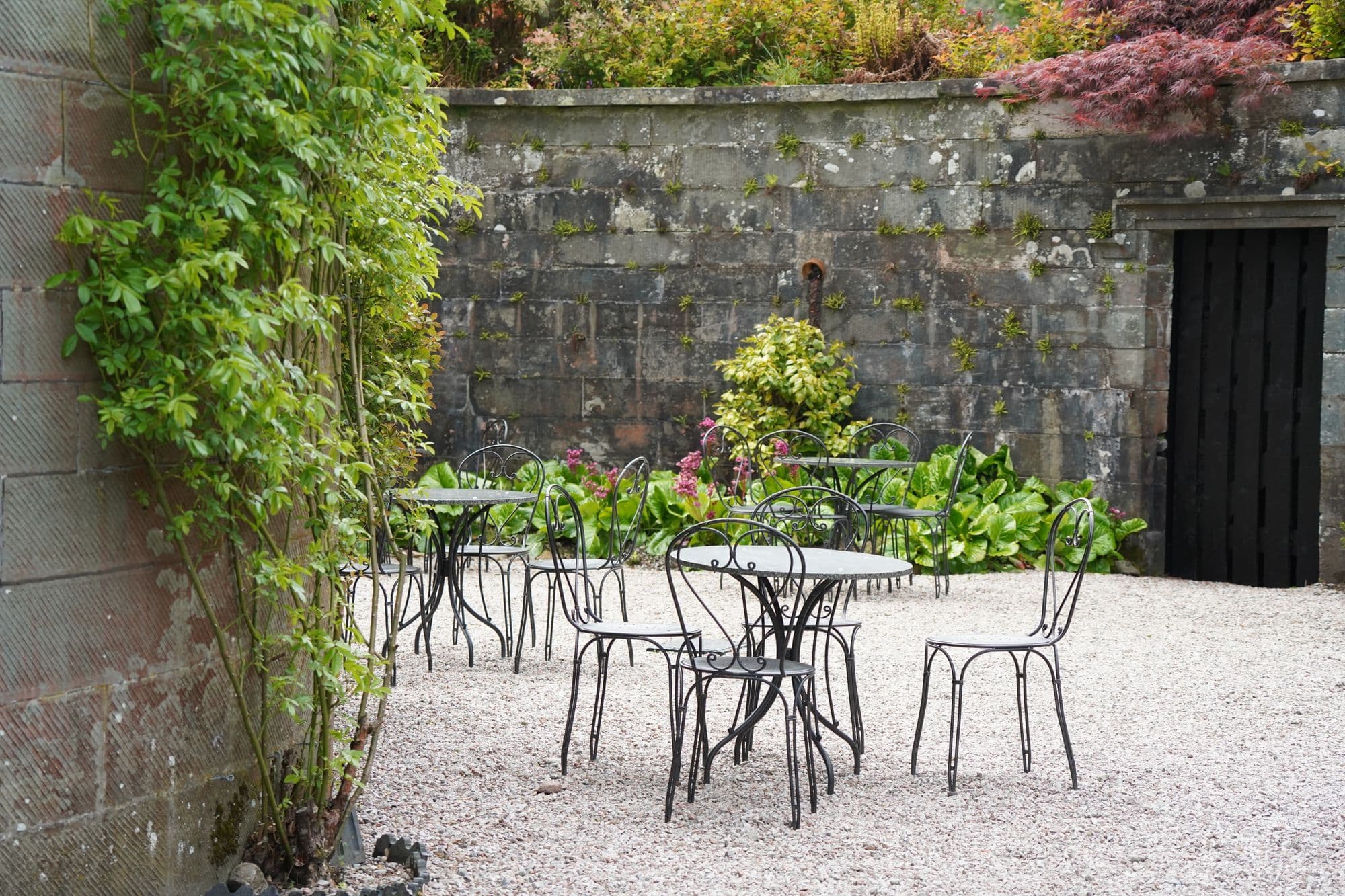 Een ommuurde tuin met grind, smeedijzeren tafeltjes en stoelen en een klimplant langs de muur
