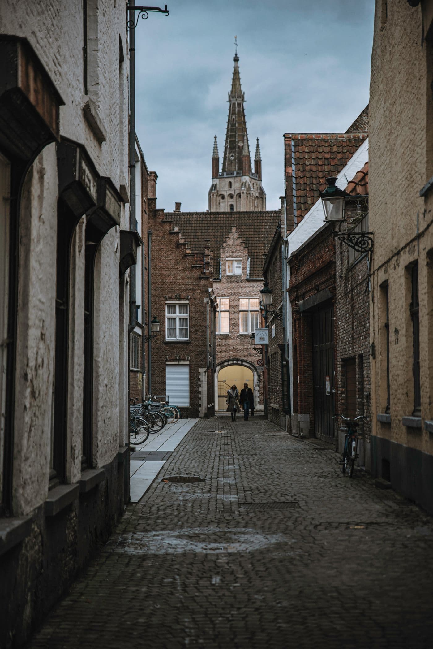 Een smal straatje met kasseien tussen oude huizen, met de toren van de Onze-Lieve-Vrouwekerk erboven