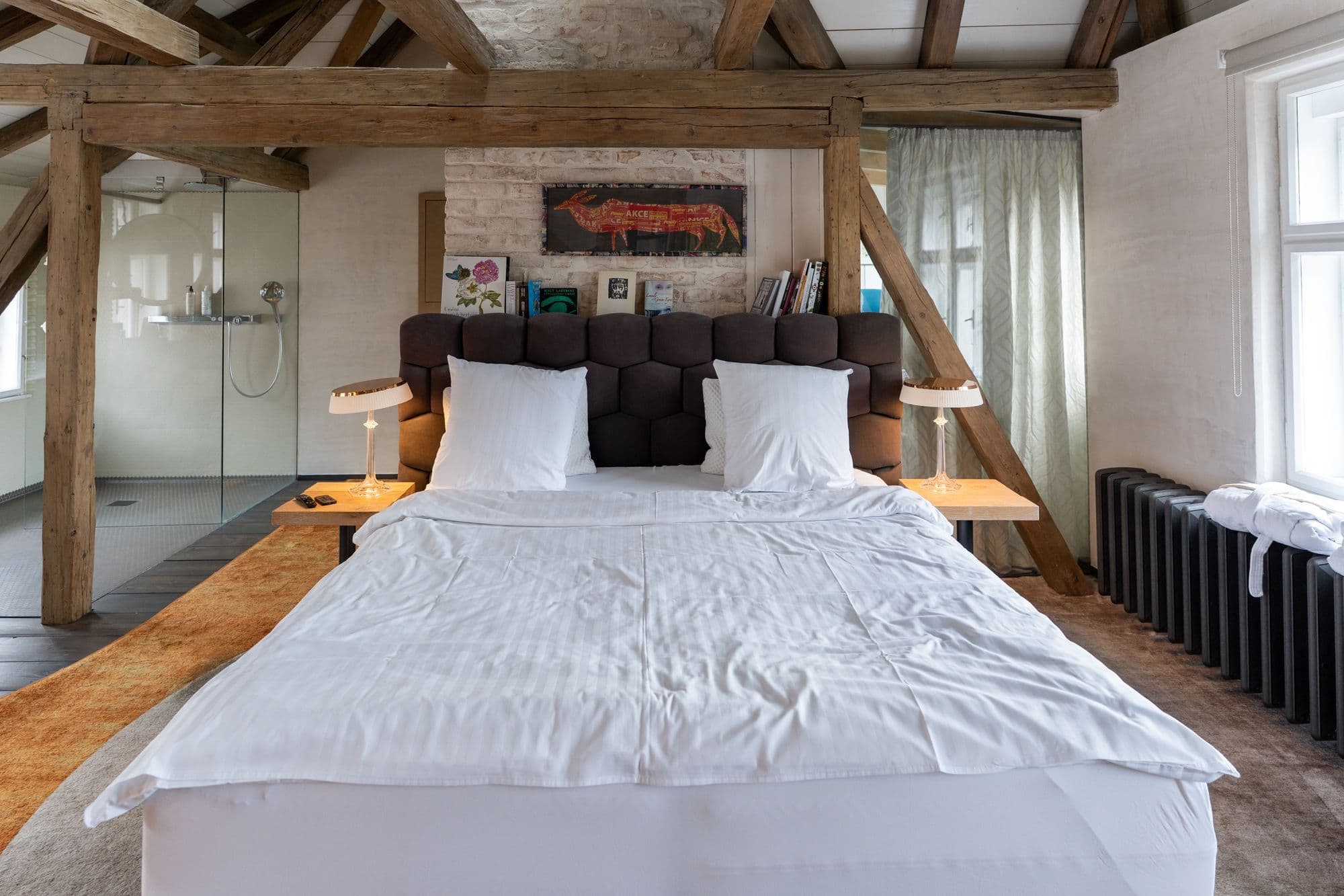 Een lichte kamer onder oude houten balken, met een tweepersoonsbed in wit linnen, een raam met vitrage en een inloopdouche achter glas