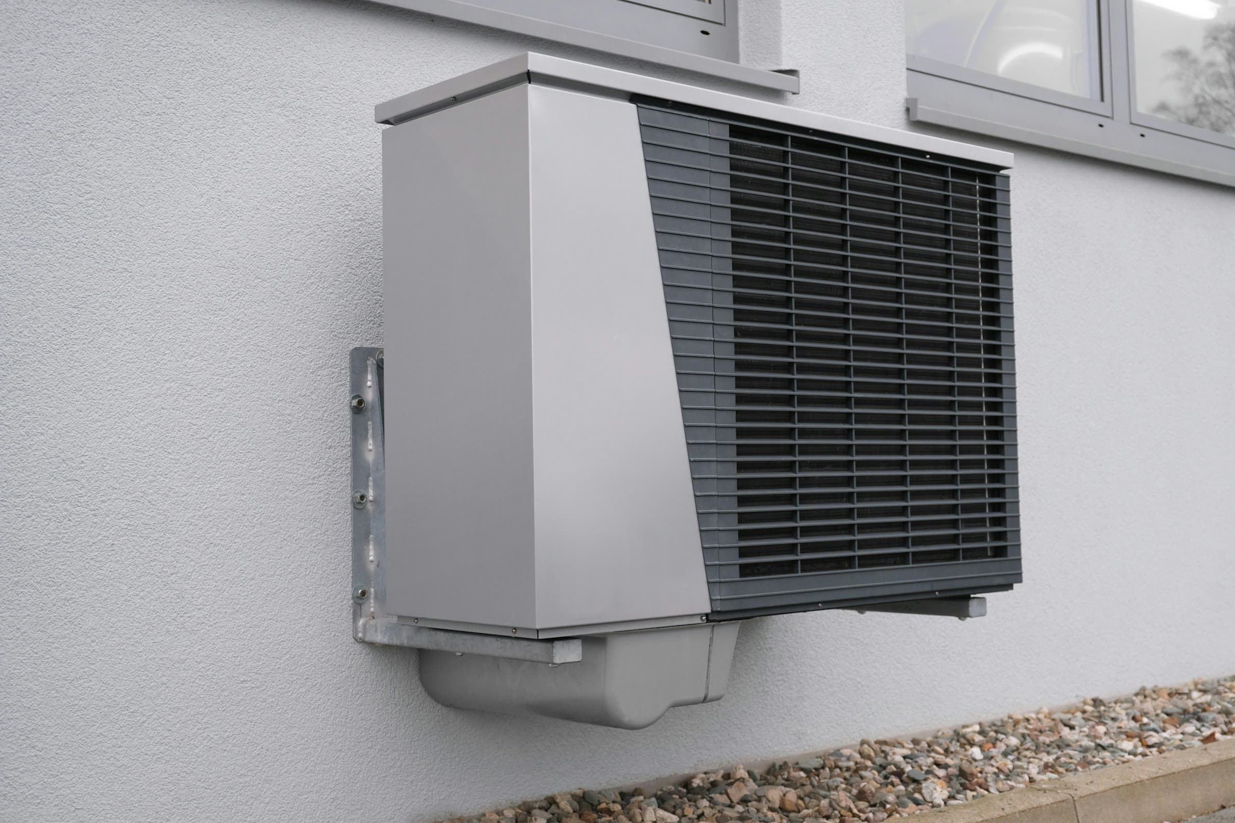 Buitenunit van een warmtepomp op een gegalvaniseerde muurbeugel tegen een witte gevel, boven een strook grind