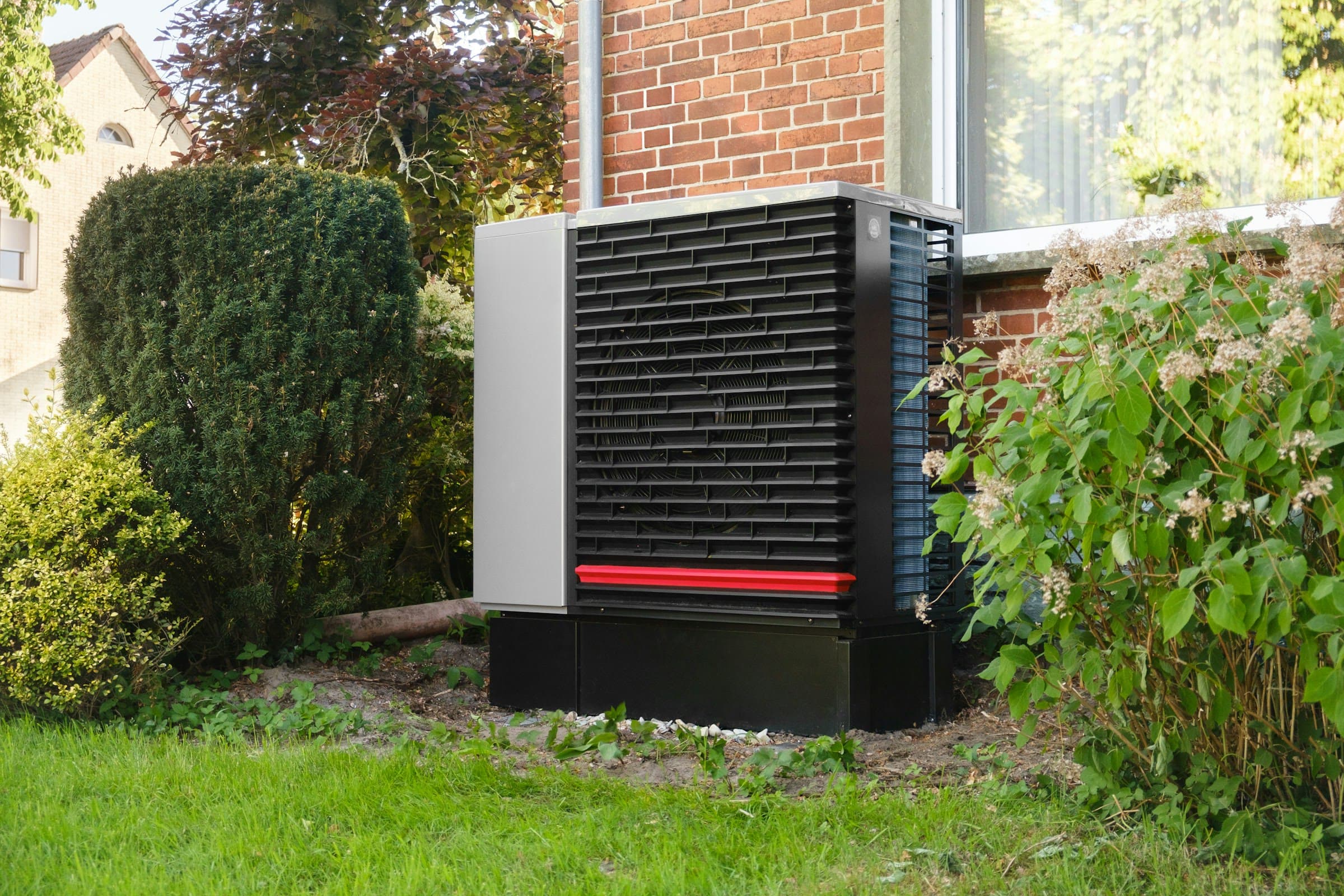 Buitenunit van een warmtepomp op een zwarte sokkel in een tuin, tussen een taxushaag en een hortensia, tegen een bakstenen gevel