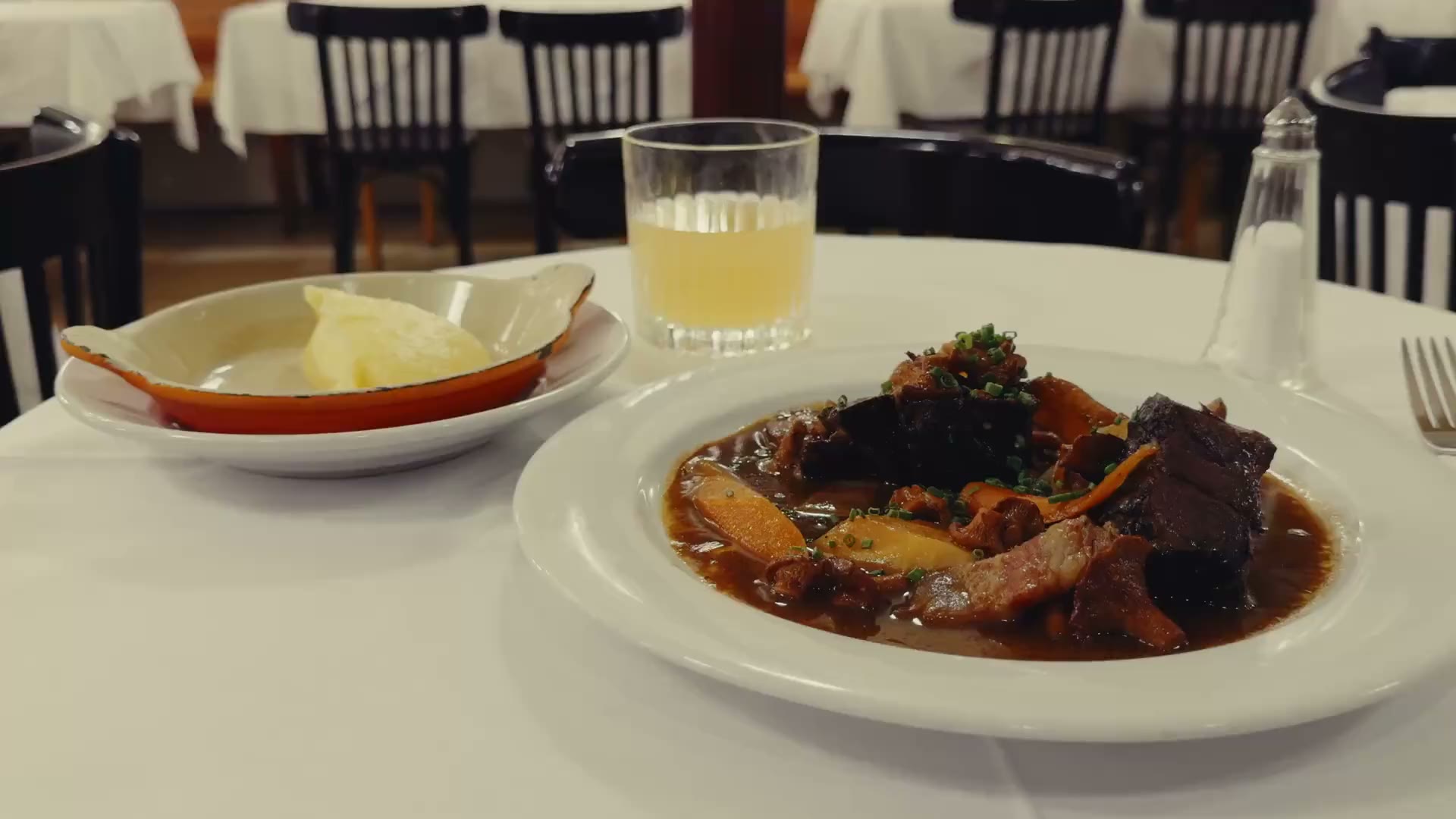 Film: een bord stoofvlees met een schaaltje puree op een wit tafellaken, achteraan de zaal met de thonetstoelen